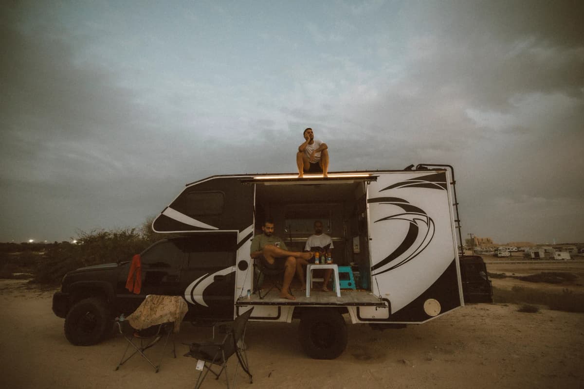 Three friends enjoying a camping trip in Dubai's desert, relaxing near a camper van.